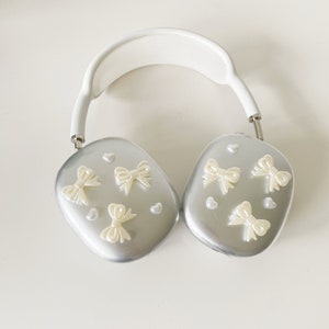 May include: White headphones with a silver finish and a white headband. The earcups are decorated with small white bows and hearts.