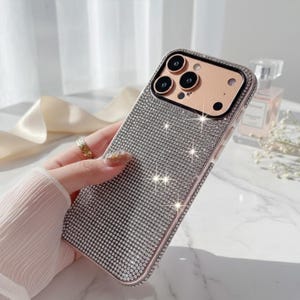 May include: A sparkling, silver rhinestone-covered phone case with a rose gold frame. The case has a camera cutout with three lenses and a flash. The phone case is held by a person.
