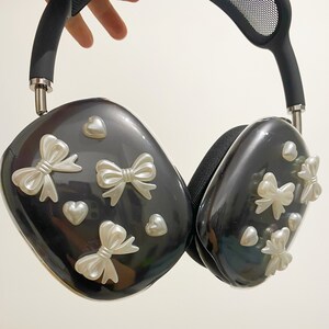 May include: Black headphones with a mesh headband and white bow accents. The headphones have a glossy finish and are decorated with small white hearts.
