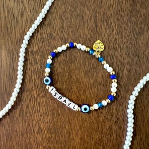 May include: A gold bracelet with white and blue beads, including evil eye charms and letter beads spelling "ISRAEL". The bracelet has a gold heart charm with the words "Love Him Above" engraved on it.