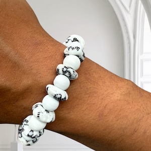 Peut inclure: Un bracelet de perles blanches et noires. Le bracelet présente un mélange de perles rondes et de forme irrégulière. Les perles ont un motif noir moucheté sur fond blanc. Le bracelet est porté au poignet d'une personne.