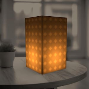 Puede incluir: Una lámpara rectangular que emite una cálida luz naranja, adornada con un patrón de diamantes repetido. La lámpara está colocada sobre una mesa redonda de madera. Una pequeña planta en maceta es visible en el fondo.