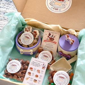 May include: A cardboard gift box containing a purple pet bowl labelled Stardust, a purple treat canister with a bone-shaped handle, a beige plastic scoop, and three clear bags of assorted pet treats. Each item features Little Companion Café branding.