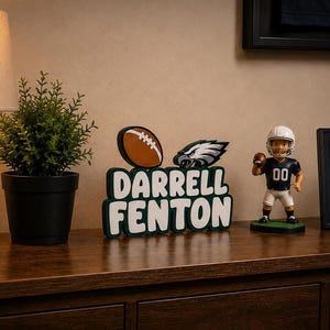 Op de afbeelding: Een gepersonaliseerd houten bord met de naam DARRELL FENTON in witte blokletters met een donkergroene omlijning, bekroond met een bruine football en het logo van de Philadelphia Eagles, naast een football-speler bobblehead-figuur.