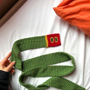 May include: A green and yellow striped knitted scarf with a red and yellow rectangular accent piece. The accent piece has two embroidered zeros. The scarf is lying on a white surface.