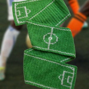 Pode incluir: Cachecol verde de crochê com um design de campo de futebol com linhas e marcações brancas. O cachecol está dobrado, mostrando o layout do campo. O fundo está desfocado, com indícios de jogadores de futebol ao fundo.