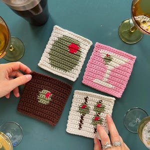 May include: Four crocheted coasters with different designs: a green olive with a red pit, a white martini glass, a brown olive with a red pit, and two green olives with red pits on sticks. The coasters are on a teal surface.
