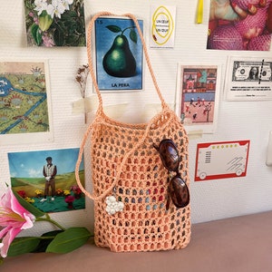 May include: A peach-coloured crocheted tote bag with a white heart charm hanging from the handle. Sunglasses are tucked inside the bag.
