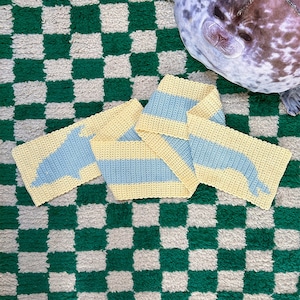 May include: A crocheted scarf with a yellow and blue striped pattern. The scarf is shaped like a dolphin and is lying on a green and white checkered rug.