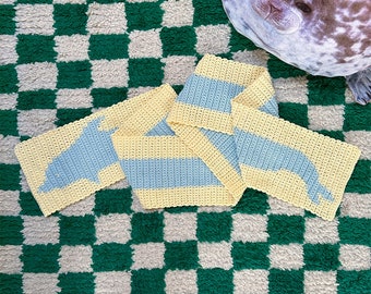 May include: A crocheted scarf with a yellow and blue striped pattern. The scarf is shaped like a dolphin and is lying on a green and white checkered rug.