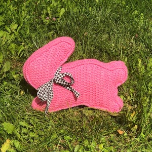 May include: A pink knitted bunny-shaped pillow with a black and white checkered bow. The plush toy is lying on a bed of green grass. The bunny has long ears and a rounded body.