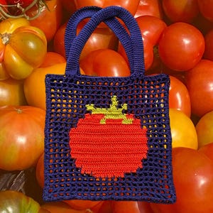 Puede incluir: Un bolso tote de crochet azul marino con un diseño de tomate rojo y tallo amarillo. El bolso tiene forma cuadrada con un patrón de malla y dos asas azul marino. El fondo está lleno de tomates maduros.