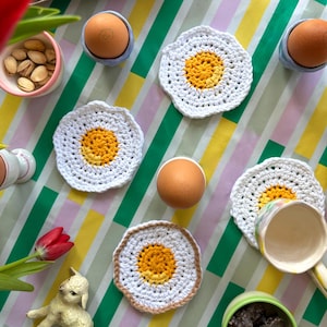 Puede incluir: Vista aérea de una mesa colorida con decoraciones de Pascua. Posavasos blancos de ganchillo en forma de huevo con centros amarillos están dispersos alrededor de huevos marrones en hueveras azules y blancas. También se ve una pequeña figura de cordero y un tulipán rojo.