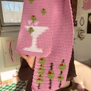 May include: A pink crocheted scarf featuring a white martini glass and green olives. The scarf also has a pattern of green olives with red centres.