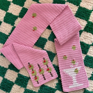 May include: A pink crocheted scarf featuring a design of green olives and a white martini glass. The scarf is laid out on a green and white chequered rug.