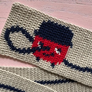 May include: A beige crocheted scarf with a red, white and blue cartoon face design. The face has a black hat and a black moustache.