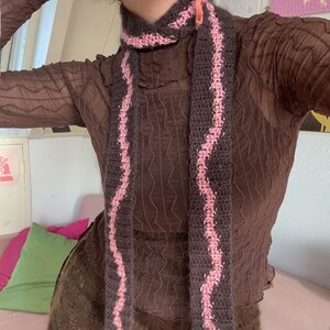 May include: A brown knitted scarf with pink stripes. The scarf is crocheted and has a wavy pattern.