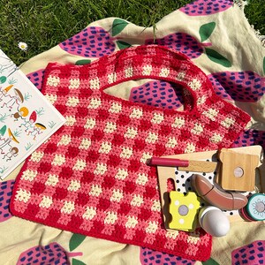 May include: A red and white crocheted picnic basket with a checkered pattern. The basket is sitting on a picnic blanket with a pink and green apple pattern.