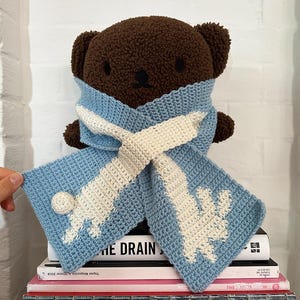 May include: A brown teddy bear wearing a light blue and white crochet scarf. The scarf has a snowflake design and a small white ball on one end. The bear is sitting on a stack of books.
