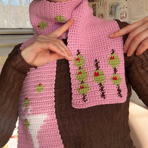 May include: A pink crocheted scarf featuring a white martini glass and green olive design. The scarf has a repeating pattern of green olive branches with red berries.