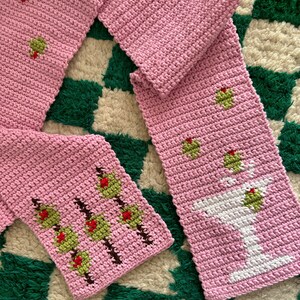 May include: A pink crocheted scarf with a green and red floral design on one end and a white crocheted martini glass on the other end.