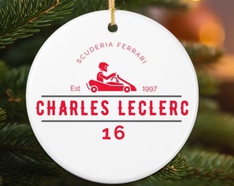 May include: A white, round ceramic ornament with a red Scuderia Ferrari logo and Charles Leclerc's name and racing number 16. The ornament is hanging from a gold string against a blurred Christmas tree background.