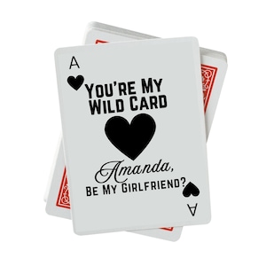 May include: White playing card with the text "YOU'RE MY WILD CARD" and "Amanda, BE MY GIRLFRIEND?" in black. The card features a black heart and the Ace of hearts symbol. A deck of cards is visible in the background.