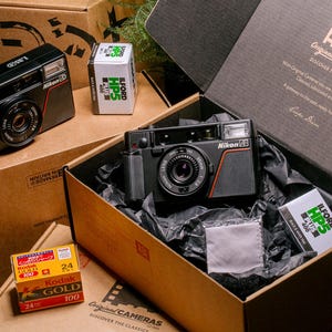 Product shot of the Nikon L35AD (Pikaichi) film camera, displayed in a cardboard "Original Cameras" packaging box with Ilford HP5 Plus film
