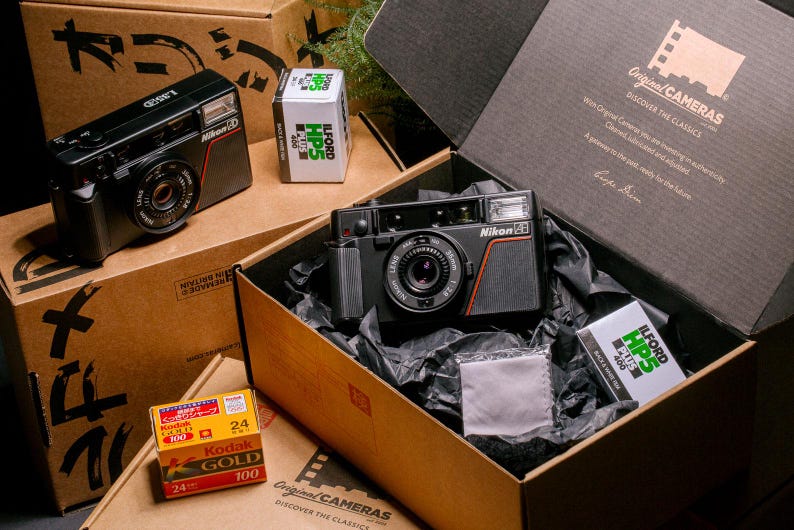 Product shot of the Nikon L35AD (Pikaichi) film camera, displayed in a cardboard "Original Cameras" packaging box with Ilford HP5 Plus film