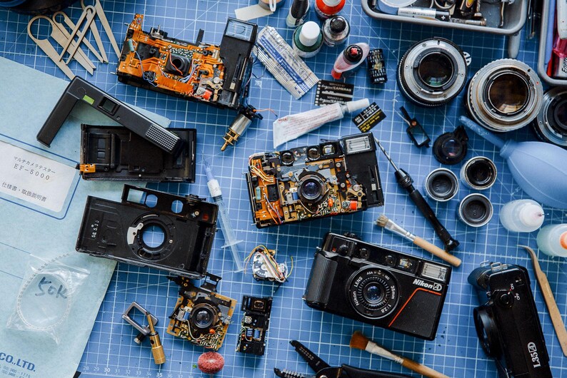 A busy Workbench with multiple disassembled L35 Nikon cameras, tools, film rolls, and various camera parts arranged together on worktop.