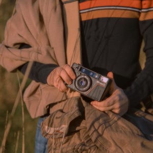 A close-up of a person holding a Nikon L35AD (Pikaichi) 35mm film camera outdoors. They have a jacket draped over their shoulder and a Domke camera bag in hand. The image captures a warm, vintage feel with natural lighting.