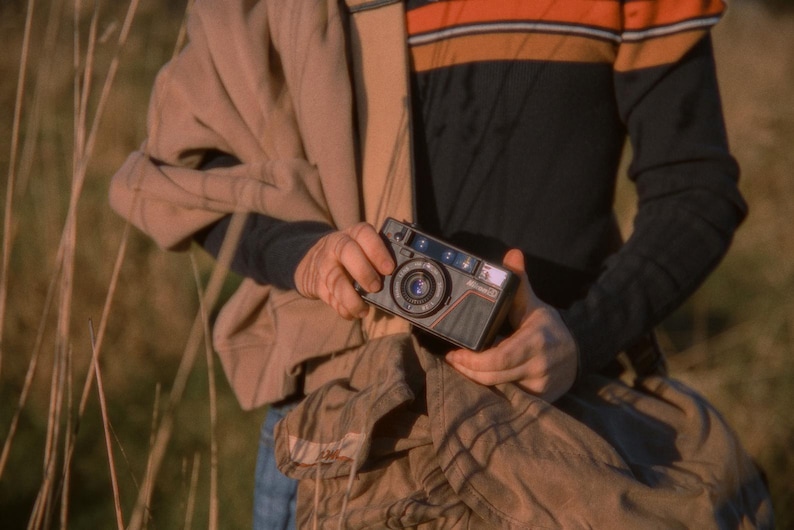 A close-up of a person holding a Nikon L35AD (Pikaichi) 35mm film camera outdoors. They have a jacket draped over their shoulder and a Domke camera bag in hand. The image captures a warm, vintage feel with natural lighting.