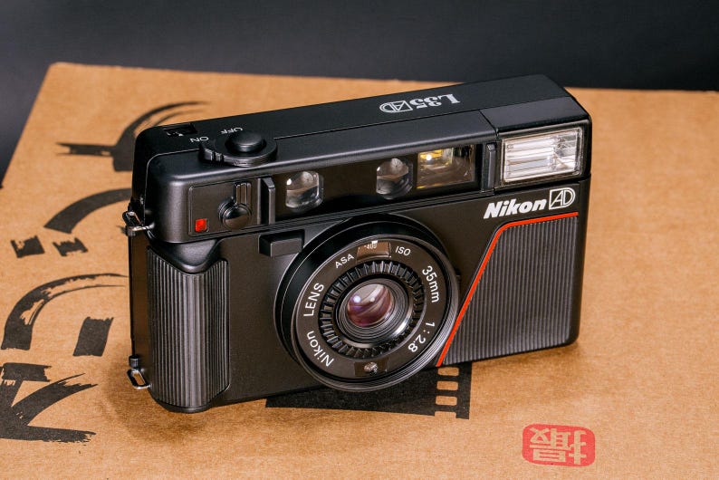 Front view of the Nikon L35AD (Pikaichi) film camera, facing above, showing the lens, graphics and built-in flash.