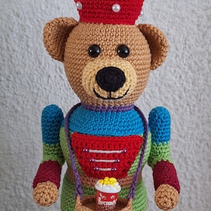 May include: A crocheted teddy bear nutcracker wearing a red hat with white popcorn and holding a basket with a small cup of popcorn that says "Popcorn".