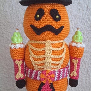 May include: A crocheted Halloween skeleton doll wearing a pink and orange hat with a black brim and a pink and orange striped belt. The doll is holding two green and white candy canes in its hands and is standing on a pink and green base.