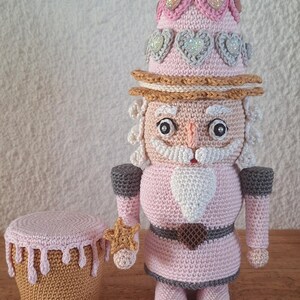 May include: A pink crocheted nutcracker doll with a white beard and a pink and grey hat decorated with sparkly hearts. The doll is holding a gold star in its right hand and is sitting on a wooden surface.