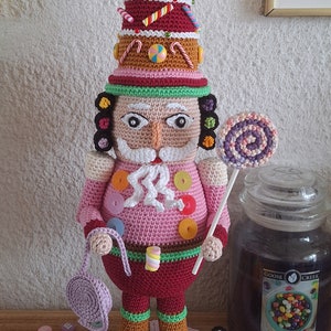 May include: A crocheted nutcracker doll holding a candy cane lollipop in one hand and a purple crocheted bag in the other. The doll is wearing a pink and red outfit with a candy-themed hat. The doll is standing on a pink and white crocheted base surrounded by colourful candy.