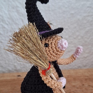 Op de afbeelding: Gehaakte heksenfiguur met een zwarte hoed en cape, die een bezem vasthoudt. De heks heeft een bruin lichaam, een roze neus en handen en een strooien bezem. Een rood lint zet de bezem vast. Een Halloween decoratie.