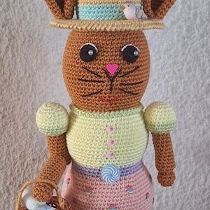 Puede incluir: Un muñeco de conejito de Pascua de crochet que lleva un sombrero con un pajarito pequeño en la parte superior. El conejito sostiene una cesta de huevos de Pascua y está de pie sobre un lecho de hierba verde con más huevos alrededor.