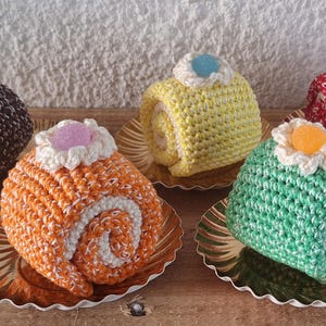 Op de afbeelding: Vijf gehaakte rolcakes op kleine gouden bordjes. De cakes zijn in verschillende kleuren, waaronder bruin, oranje, geel, groen en rood. Elke cake heeft een klein decoratief stuk stof erop.