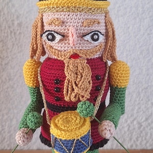 May include: A crocheted nutcracker doll holding drumsticks and a drum. The doll is wearing a red and black uniform with a gold trim and a black hat with a green holly sprig. The doll is standing on a yellow base.