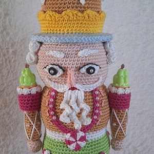 May include: A crocheted gingerbread nutcracker doll wearing a hat with white icing and blue sprinkles, a green and pink candy cane dress, and holding two green and white candy canes.