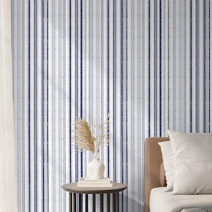 May include: A bedroom with a white and blue striped wallpaper. A bed with a white duvet and a brown headboard is in the foreground. A small table with a vase of dried flowers sits next to the bed.
