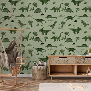 Op de afbeelding: Een groene kinderkamerbehang met een herhalend patroon van dinosaurussen en palmbomen. Het behang bevindt zich in een kamer met een schommelpaard, een hangende hangmat en een houten plank.