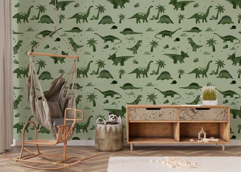 Op de afbeelding: Een groene kinderkamerbehang met een herhalend patroon van dinosaurussen en palmbomen. Het behang bevindt zich in een kamer met een schommelpaard, een hangende hangmat en een houten plank.