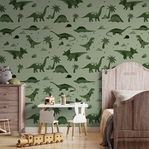 Op de afbeelding: Een groen-wit behang met een herhalend patroon van dinosaurussen en palmbomen. Het behang is in een kinderkamer met een houten bed, een commode en speelgoed.