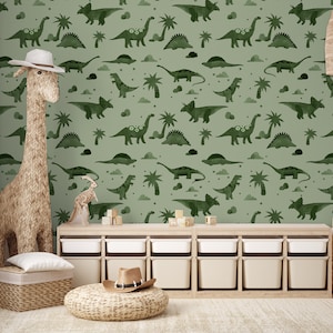Op de afbeelding: Een groene en witte dinosaurus-thema wallpaper met een herhalend patroon van dinosaurussen, palmbomen en sterren. De wallpaper is in een kinderkamer met een houten speelgoedrek en een giraffen speelgoed.