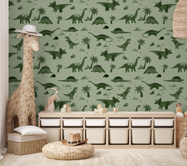 Op de afbeelding: Een groene en witte dinosaurus-thema wallpaper met een herhalend patroon van dinosaurussen, palmbomen en sterren. De wallpaper is in een kinderkamer met een houten speelgoedrek en een giraffen speelgoed.