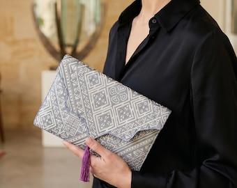May include: A rectangular clutch with a geometric pattern in shades of gray and white, accented with a purple tassel. The clutch is held against a black shirt.