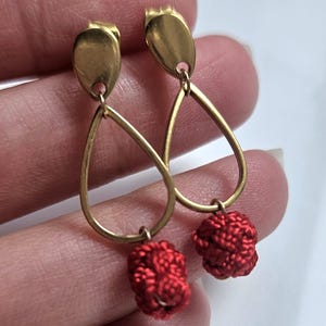 Puede incluir: Un par de pendientes de aro dorados con borlas de tela roja. Los pendientes tienen un diseño en forma de lágrima con una pequeña bola en la parte inferior.